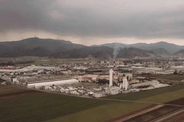 Aerial photo of a factory sitting between the green fields. Visible smoke coming out from a smokestack. Factory in the nature on a dull day, concept of dirty factory in nature.