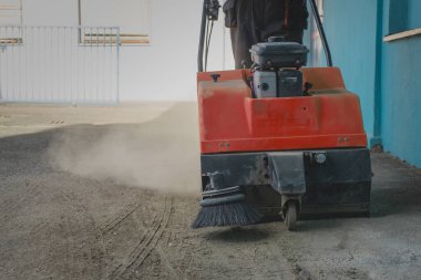 Birisi, kirli asfalt zemini üzerinde turuncu benzinle çalışan bir endüstriyel süpürgeyi enkaz ve kumdan temizlemeye çalışıyor.
