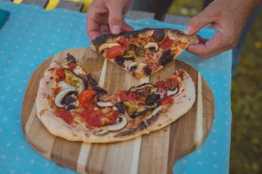 Ev yapımı pizza taze pişmiş ve bahçedeki bir kırsal masada ahşap bir tabakta servis ediliyor. Bir pizza için güzel bir ortam. Bir insanın elleri bir dilim alırken görülüyor..