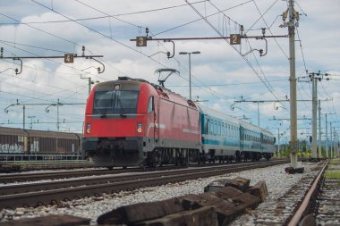 Banliyö istasyonunda kırmızı motorlu modern elektrikli tren. Bulutlu bir günde, banliyö ortamında şehre doğru koşan yolcu treni.
