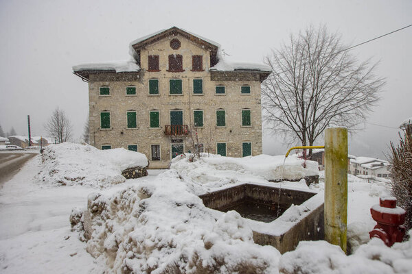 Italian village square in snow or during heavy snowfall with visible fountain as the centre of the village. Water still flowing even in winter time. Majestic town house in the background.