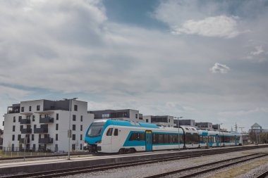 Modern bir konut veya yerleşim yerinin önündeki bir istasyonda modern beyaz ve mavi yolcu treni. Şehir blokları ve tren.