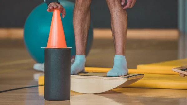 Male trainer seen Touching a cone while on wooden balance board. Different balance sports equipment is seen around in a gym indoors.