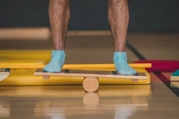 Front view of male trainer seen using a wooden balance board. Different balance sports equipment is seen around in a gym indoors.