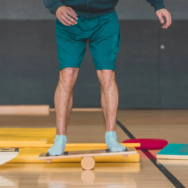 Front view of male trainer seen using a wooden balance board. Different balance sports equipment is seen around in a gym indoors.