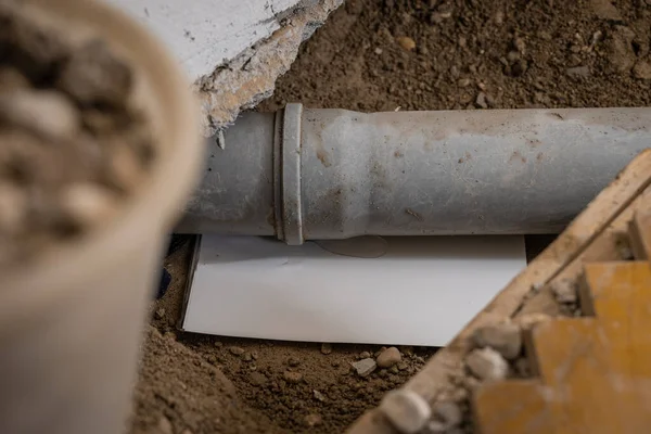 Leaking plastic sewage pipe in an old apartment, visible open hole with removed parquet, sand and buckets around, exposed drain pipe in the ground.