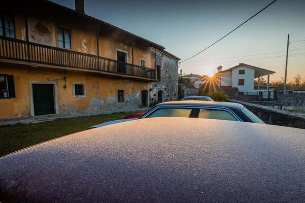 Frost-covered classic car roof in foreground at sunrise, with rustic yellow stone house, green doors, balcony, and village buildings bathed in golden light.