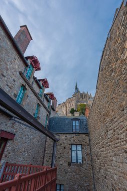 Mont Saint-Michel, Fransa 'da dar bir sokak. Yarı ahşap duvarlar, klasik fener ve taş cepheler, bulutlu bir gökyüzüne karşı manastırın altın kulesini andırıyor..