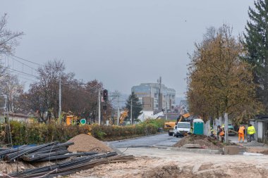 Ljubljana 'daki Goriska Caddesi' ndeki yol yapımında, arka planda binalar ve sonbahar ağaçları bulunan sisli bir şehir yolu boyunca işçiler, makineler ve malzemeler görülüyor..