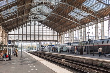 Tarihi La Rochelle tren istasyonunun geniş bir manzarası var. Devasa kemerli cam ve çelik çatının egemenliğinde. Bir tren peronda bekliyor. Yolcular rayların yakınında dolanıyor..