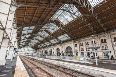 Tarihi La Rochelle tren istasyonunun geniş bir manzarası var. Devasa kemerli cam ve çelik çatının egemenliğinde. Bir tren peronda bekliyor. Yolcular rayların yakınında dolanıyor..