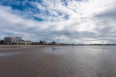 Royan Sahili 'nin panoramik çekimlerinde dağınık bulutlarla dolu dramatik bir gökyüzü altında geniş bir kum ve sakin bir su vardır. Modern binalar uzak kıyılarda..
