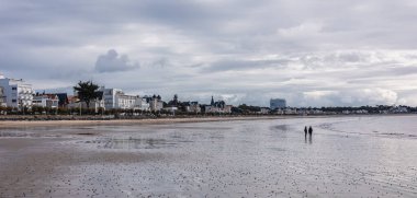 Royan Sahili 'nin panoramik çekimlerinde dağınık bulutlarla dolu dramatik bir gökyüzü altında geniş bir kum ve sakin bir su vardır. Modern binalar uzak kıyılarda..