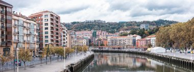 Durgun suya yansıyan renkli binalarla Bilbao nehir kıyısının geniş manzarası. Şehir, çarpıcı, bulutlu bir gökyüzünün altında, kentsel ve doğal elementleri gözler önüne seren ağaçlık bir yamaçtan yükseliyor..