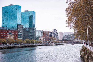 Bilbao 'daki Nervi n Nehri' nin geniş bir manzarası, şehrin modern cam kuleleri, La Salve köprüsü ve parlak bulutlu gökyüzü altında ağaçlarla kaplı bir kıyı..