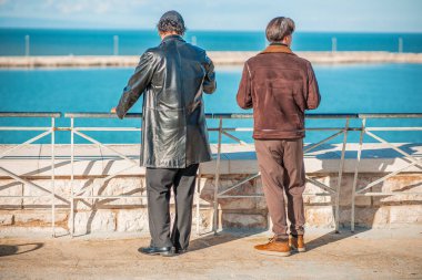 İki adam birlikte rıhtımda duruyorlar, parlak mavi denize bakarken sakin bir anı paylaşıyorlar. Yakınlıkları ve rahat duruşları, sahneyi, deniz kenarında huzurlu bir günün tadını çıkaran bir çiftin sıcaklığına dönüştürüyor..