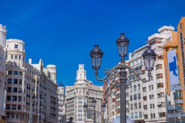 Bu resim Valencia 'daki Plaza del Ayuntamiento' yu gösteriyor, antik bir sokak lambasından görülüyor. Arka plan, büyük neoklasik cepheleri ve parlak mavi Akdeniz gökyüzünü vurguluyor.