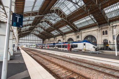 Bu geniş açılı çekim La Rochelle tren istasyonunu yakalar. Mavi ve gümüş renkli bir tren, karmaşık taş mimari, sayısız paralel raylar ve bulutlu gökyüzü ile birlikte platformda oturur..