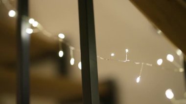 İnce gümüş bir tel üzerinde, koyu dikey sütunlara sarılı narin, ılık, beyaz peri ışıkları parıldıyor. Sığ alan derinliği tatil dekoru için rüya gibi, bokeh dolu bir atmosfer yaratıyor..