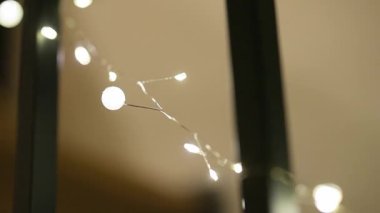 İnce gümüş bir tel üzerinde, koyu dikey sütunlara sarılı narin, ılık, beyaz peri ışıkları parıldıyor. Sığ alan derinliği tatil dekoru için rüya gibi, bokeh dolu bir atmosfer yaratıyor..