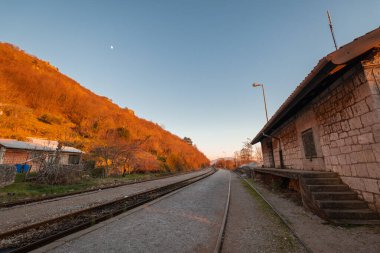 Altın saat buzet tren istasyonu, taş binalar ve kavisli rayların yanında antika bir platform. Sıcak gün batımı ışığı yakınlardaki evleri ve kayalık tepeleri yıkıyor, sakin, kırsal bir Istrian manzarası yaratıyor..