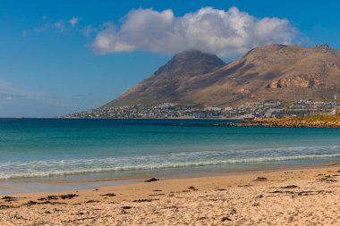 Simons Town, Güney Afrika 'nın panoramik manzarası: kıvrımlı bir kıyı şeridi boyunca kurulmuş renkli kıyı evleri ve sahte körfez suları. Masa dağları, dağılmış bulutlarla parlak mavi bir gökyüzünün altında görünür..