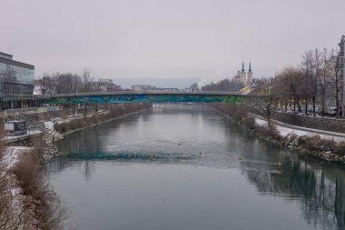 Avusturya 'nın Villach kentindeki Drava Nehri' nin sakin bir kış manzarası, boyalı yaya köprüsü, karlı nehir kıyıları, deniz yansımaları, ve uzakta ikiz kuleleri olan bir kilise..