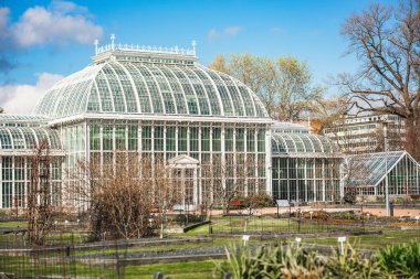 Helsinki Üniversitesi Botanik Bahçesi 'nin zarif, beyaz Palm House' u mavi gökyüzüne karşı parlak bir şekilde duruyor. Karmaşık 1889 cam mimarisi çakıl yollarına ve baharda uyanan bahçe yataklarına bakıyor..