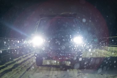 Gece karlı bir yolda parlak farları olan siyah bir minibüs duruyor. Kar taneleri ön planda, parıltının aydınlattığı bir bokeh etkisi yaratır. Sahne soğuk, sinematik ve atmosferik..