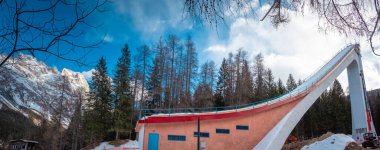 İtalya, Cortina d Ampezzo 'da kayak rampası üzerinde yükselen kayak rampası dramatik beton yapısı, mavi gökyüzü, kış ışığı ve çarpıcı bir dağ sporu sahnesi yaratan yüksek çam ağaçları..