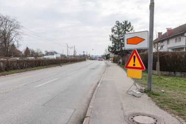 Bir Sloven yol tabelası, turuncu sapma oku ve bir üçgen uyarı levhasıyla eşleştirilmiş Tam Yol Kapanışı konusunda uyarıyor. Bulutlu bir gökyüzünün altında sokaklar temiz ve boş..