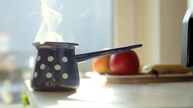 Güneşli mutfak sahnesi, üzerinde beyaz benekli küçük mavi bir kahve demliğini ahşap bir tahtanın üzerinde buharlaşırken gösteriyor. Seramik kupalar ve çaydanlık yakınlarda oturur, sıcak, sıcak bir sabah kahvesi anını yakalar..