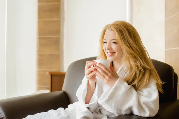 Hermosa chica rubia en una túnica blanca bebiendo de tazas y sm —  Fotos de Stock