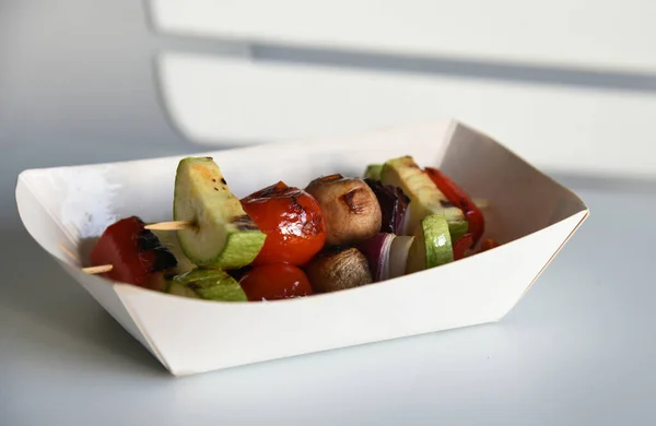 Delicious healthy meal salad with grilled vegetable, grilled tomato, grilled mushrooms, grilled onions and grilled zucchini on wooden stick. Grilled vegetables salad in white cardboard box on simple white table and blur background.