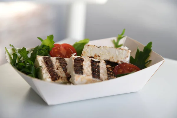 Delicious healthy meal salad with grilled cheese, fresh tomato and arugula. Salad in white cardboard box on simple white table and blur background.