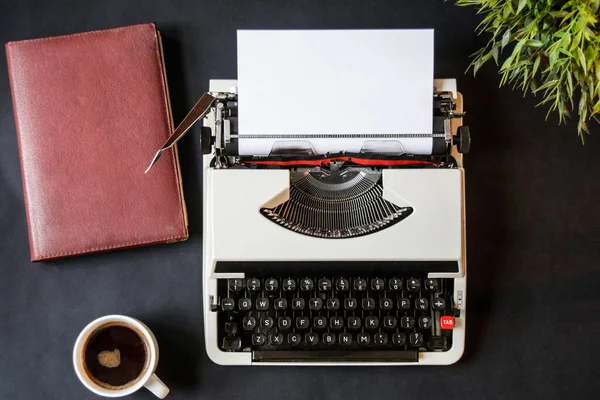 Typewriter with paper in it, artificial plant decoration, retro leather notebook and coffee cup on the dark background. Beautiful retro working space closeup. Empty space on paper to write down something.