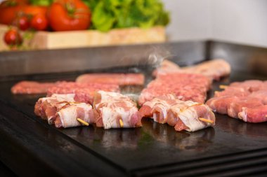 Raw meat on wooden chopsticks on hot grill with smoke. Vegetables in background