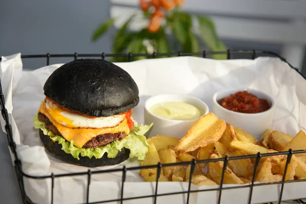 Modern meal with black burger , egg, salad, cheese and meat on white background and napkins. Fries and two kinds of sauce in white porcelain bowls. Plant of small orange pepper in background