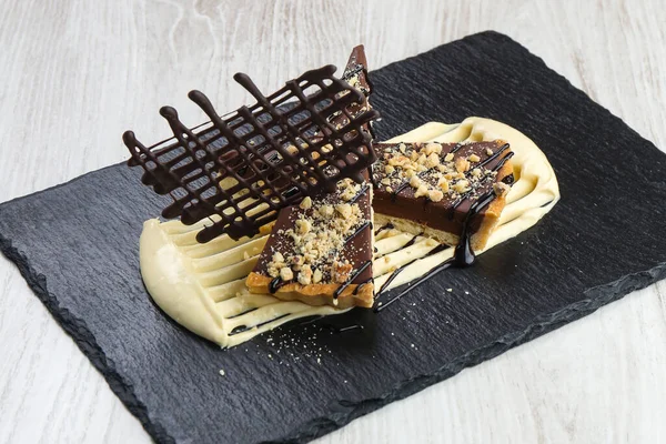 Chocolate tart cake on black marble plate in restaurant. Decoration from chocolate peaces with almond and peanuts.
