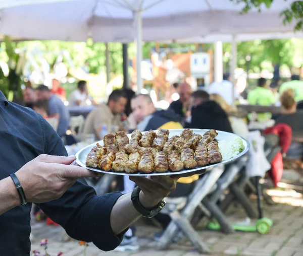 Big plate full of grilled meat. Cevapi on metal plate. Waiter holding plate full of fresh grilled meat. People eating  in restaurant in background. Blur. Masculine male hands. Empti copy space