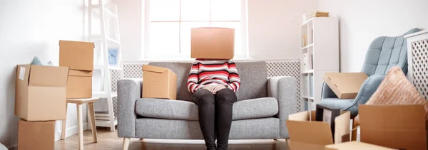 Femme fatiguée avec une boîte en carton sur la tête assise sur le canapé dans sa nouvelle maison. — Photo