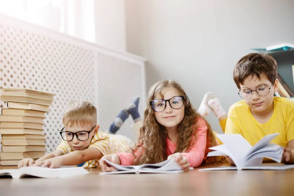 Quatre enfants mignons dans des lunettes allongées sur le sol à l'intérieur avec des livres — Photo