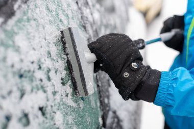 Biri buz kazıyıcı ile arabanın camlarını kazıyarak temizliyor. Yakın plan, eller kış eldivenlerinde..