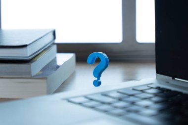 Dizüstü bilgisayarla istiflenmiş kitaplar arasına yerleştirilmiş küçük mavi bir soru işareti. Soruşturma, araştırma, eğitim, teknik destek, çevrimiçi öğrenim, kafa karıştırıcı kavramlar.
