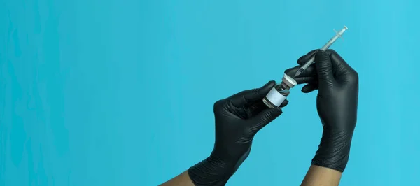 Mock-up of a bottle with a vaccine, medicine or serum with a syringe in the hands of a doctor in black medical gloves, isolated on a blue background. Beauty shot.banner
