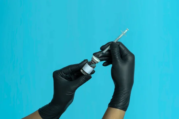 Mock-up of a bottle with a vaccine, medicine or serum with a syringe in the hands of a doctor in black medical gloves, isolated on a blue background. Beauty shot