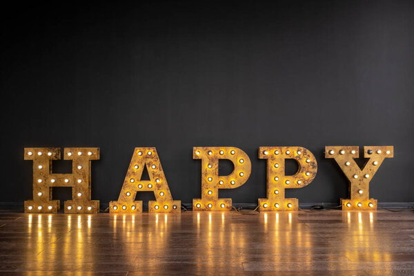Слово happy выполнено из больших желтых металлических букв с перламутровыми лампочками на полу на фоне стены