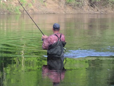 Tanımlanamayan, tulumlu ve şapkalı olgun bir erkek nehirde uzun ince bir oltayla balık tutarken gündüz vakti 
