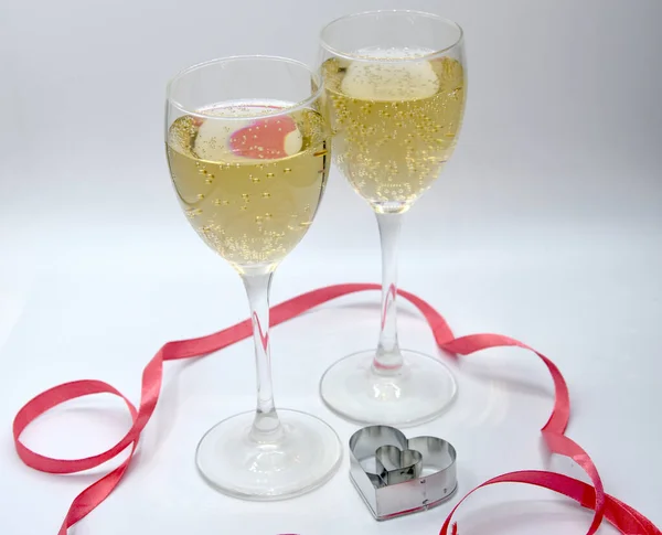 Şeffaf bardaklarda açık sarı şampanya kadehlerinin seçici odak noktası ve kalp şeklinde küçük metalik kurabiye kesici ve beyaz arka plandaki lambalar ışığında parlak pembe kurdele. 