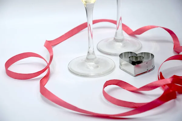 Small gray metallic cookie cutter in form of heart and transparent stems of glasses surrounded by long thin snaking vibrant pink ribbon on white table in light of lamps